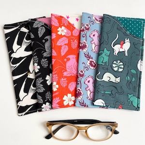 Peut inclure: Plusieurs étuis à lunettes en tissu coloré avec différents motifs, notamment des oiseaux, des chats et des motifs floraux. Les étuis sont disposés verticalement, avec une paire de lunettes en bas. Les lunettes ont une monture marron.