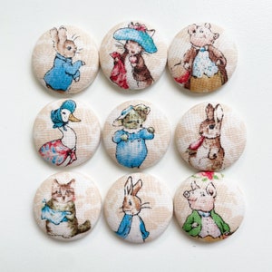 May include: Nine round fabric buttons featuring characters from the children's book, Peter Rabbit. The buttons are beige with a white lace pattern and feature characters like Peter Rabbit, Benjamin Bunny, Jemima Puddle-Duck, Mrs. Tiggy-Winkle, and more.