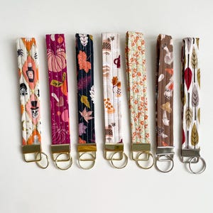 May include: Seven fabric key fobs with autumn-themed patterns. Designs include pumpkins, leaves, and coffee cups. Each fob is attached to a gold or silver-coloured metal ring. The key fobs are made of quilted fabric.