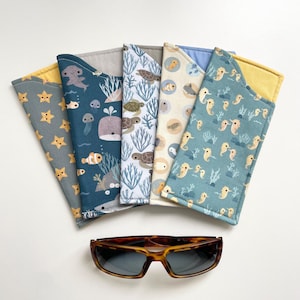 May include: Five fabric eyeglass cases with marine life patterns, including stars, whales, turtles, and seahorses. Tortoiseshell sunglasses are below the cases. The cases have a soft, protective design, ideal for storing glasses.