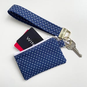 Op de afbeelding: Marineblauwe polkadot polsband met een bijpassend etui met ritssluiting. De etui bevat een zwarte kaart met het woord "WIN" en een rode kaart. Een goudkleurige metalen sluiting verbindt de polsband met de etui en een sleutel.
