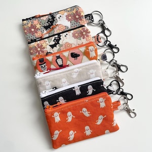 May include: A collection of six rectangular fabric zip pouches with attached metal key ring clips. The pouches feature various patterns including white ghosts, flowers, stars, bats, skulls, and potion bottles in orange, black, and cream colours.