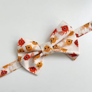 Könnte beinhalten: Ein weißer Fliege mit einem Muster aus orangefarbenen und roten Goldfischen. Die Fliege ist aus Stoff und hat ein schlichtes Design.