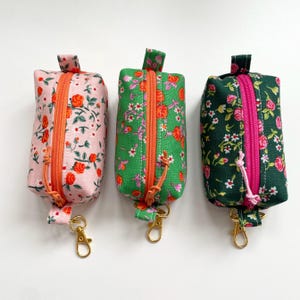 May include: Three small, rectangular pouches with floral patterns. Each pouch has a zip closure and a gold-coloured clasp. The pouch on the left is pink with orange roses, the middle pouch is green with orange roses, and the right pouch is dark green with pink roses.