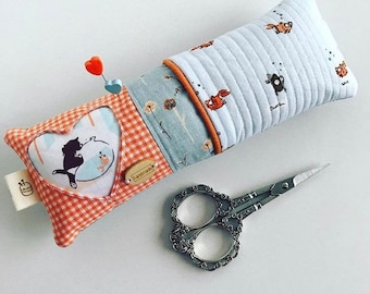 May include: A handmade pincushion with a patchwork design featuring a heart with a cat and fish illustration, a blue and white fabric with a floral pattern, and a white fabric with a repeating pattern of fish. The pincushion is filled with stuffing and has a wooden tag that says "Handmade". A pair of silver scissors with a floral design is also pictured.