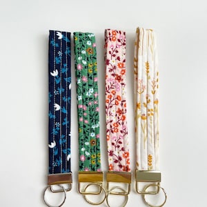 May include: Four fabric wrist keychains with floral patterns in navy, green, pink, and cream. Each keychain has a gold-coloured metal clasp and ring. The keychains are quilted with vertical stitching.