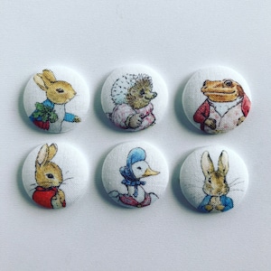 May include: Six round fabric buttons featuring characters from the children's book, Peter Rabbit. The buttons feature Peter Rabbit, Benjamin Bunny, Mrs. Tiggy-Winkle, Mr. McGregor, Jemima Puddle-Duck, and a hedgehog.