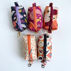 May include: Five small, rectangular zippered pouches with keychains. The pouches feature various autumn-themed patterns, including leaves, pumpkins, and floral designs. The zippers are in colours like purple, orange, and burgundy. The pouches are likely for carrying small items.