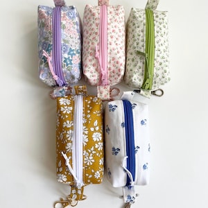 May include: Six small, rectangular zippered pouches with floral patterns. The pouches feature a variety of colours, including purple, pink, green, yellow, and white. Each pouch has a matching coloured zip and a keychain.