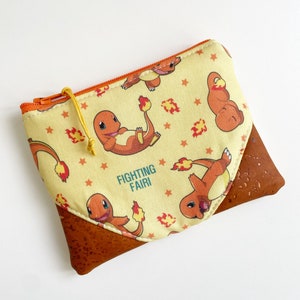 May include: A small yellow zippered pouch with a brown cork bottom. The pouch is decorated with a repeating pattern of orange cartoon characters with flames and stars. The text "FIGHTING FAIRI" is printed on the pouch.