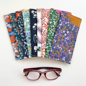 May include: A collection of ten fabric spectacle cases arranged in a fan shape, each featuring a distinct floral or botanical pattern in various colours including blue, green, yellow, and pink, positioned above a pair of dark red plastic spectacles.