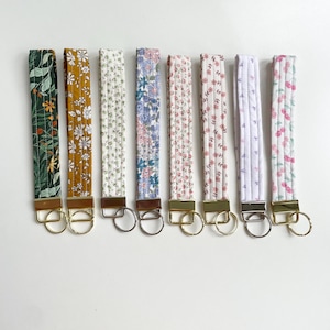 Könnte beinhalten: Eine Sammlung von acht Schlüsselanhängern für das Handgelenk aus Stoff mit verschiedenen floralen Mustern. Die Schlüsselanhänger haben gold- oder silberfarbene Hardware und eine Schlaufe zum Befestigen von Schlüsseln. Die Muster umfassen grüne Blätter, gelbe Gänseblümchen und rosa Kirschen.