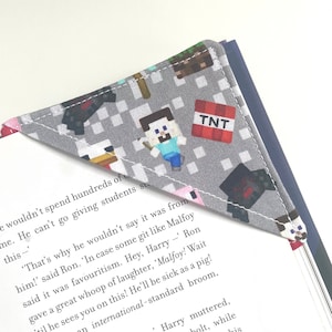 May include: A grey and white fabric bookmark with a Minecraft theme. The bookmark features a variety of Minecraft characters and items, including Steve, a creeper, a pig, a chicken, and TNT. The bookmark is shaped like a triangle and has a white border.