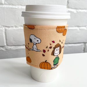 Peut inclure: Manchon de tasse à café beige avec Snoopy, Charlie Brown, des citrouilles et des feuilles d'automne. Le manchon est enroulé autour d'une tasse à café en papier blanche avec un couvercle blanc. Le design est sur le thème de l'automne.