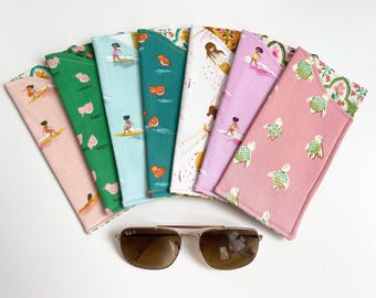 May include: A collection of colorful fabric sunglass cases with various patterns, including surfers, birds, and turtles. A pair of Ray-Ban sunglasses with a gold frame and brown lenses rests in front of the cases.