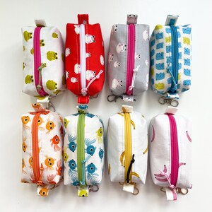 May include: Eight small, rectangular fabric pouches with zippers and keyrings. The pouches feature various patterns, including frogs, sheep, clouds, goldfish, and bananas. Each pouch has a different coloured zip, such as pink, blue, and yellow.