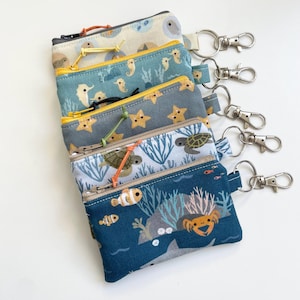 Op de afbeelding: Een stapel van vijf rechthoekige stoffen etuis met ritssluiting en bevestigde metalen karabijnhaakjes. Elk etui heeft een andere print met een zeethema, waaronder zeepaardjes, zeesterren, zeeschildpadden, anemoonvissen en krabben in blauw en wit.