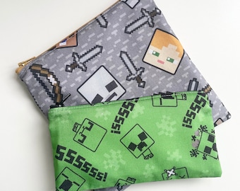 May include: Two zippered pouches with Minecraft themed designs. The larger pouch is gray with a repeating pattern of swords, bows and arrows, and characters from the game. The smaller pouch is green with a repeating pattern of Creeper faces and the text "ssssss!" in black.
