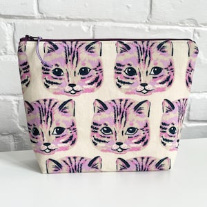 Peut inclure: Une pochette zippée blanche avec un imprimé de chat rose et noir. Les chats ont des rayures roses et noires et regardent directement le spectateur.