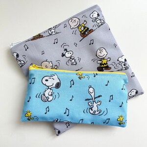 May include: Two fabric pencil cases with a Peanuts theme. The larger case is gray with black and white illustrations of Snoopy and Charlie Brown. The smaller case is blue with black and white illustrations of Snoopy. Both cases have a yellow zipper.