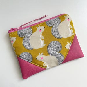 May include: A small yellow zippered pouch with a pink leather flap. The pouch is decorated with a repeating pattern of squirrels with pink cheeks.