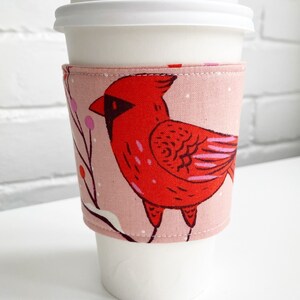 Peut inclure: Un manchon de tasse à café rose et rouge avec un motif d'oiseau cardinal. Le manchon est en tissu et a une couture cousue autour du bord supérieur.