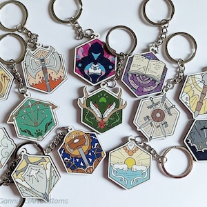 Op de afbeelding: Een verzameling zeshoekige sleutelhangers met verschillende fantasiethema's. Elke sleutelhanger heeft een andere illustratie, waaronder een luit, een bijl, een boek en een schild, met zilverkleurige sleutelringen en kettingen. Op een witte achtergrond.