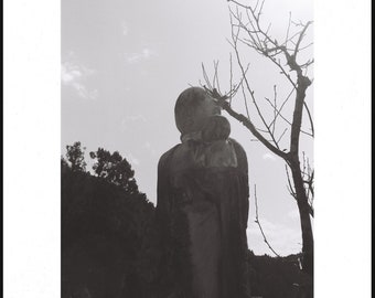 May include: A black and white photograph of a stone statue of a person holding a child. The statue is in front of a tree with bare branches and a cloudy sky.