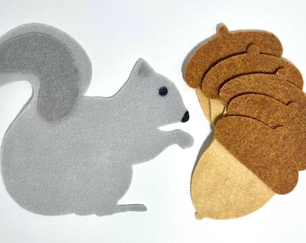 May include: A gray felt squirrel with a black nose and eye details faces a cluster of brown felt acorns with tan bases. The squirrel is positioned to the left, with the acorns on the right against a white background.