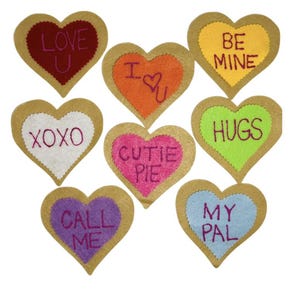 May include: Nine heart-shaped felt decorations with scalloped edges. Each heart has a different color and a message embroidered in a contrasting color, including "LOVE U," "I ♡ U," "BE MINE," "XOXO," "CUTIE PIE," "HUGS," "CALL ME," and "MY PAL."