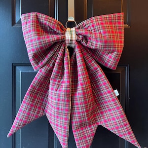 May include: A large fabric bow featuring a red, green, and yellow plaid pattern. The bow is secured with a central band of white, green, and gold checkered fabric and hangs from a silver-toned metal hook attached to a dark door.