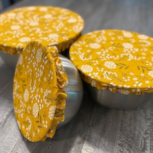 May include: Three stainless steel bowls with yellow floral fabric bowl covers. The covers have a white floral pattern on a yellow background. The bowls are on a gray surface.