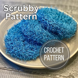 May include: Two blue crocheted scrubbies are displayed in a white ceramic dish. The scrubbies are made of a textured, looped yarn. Text on the image reads "Scrubby Pattern" and "Crochet Pattern". The background is a grey wooden surface.