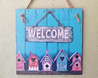 Könnte beinhalten: Ein türkisfarbenes Holzschild mit dem Wort "WELCOME" in weißen Buchstaben. Das Schild ist mit einer Reihe bunter Vogelhäuschen mit Herzen und einem Seil zum Aufhängen verziert.