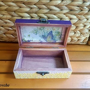 Puede incluir: Una caja de madera con una tapa morada y un dise&ntilde;o floral en el interior. La caja tiene un dise&ntilde;o de mariposa en el interior de la tapa y un dise&ntilde;o floral en el fondo. La caja est&aacute; abierta y vac&iacute;a.