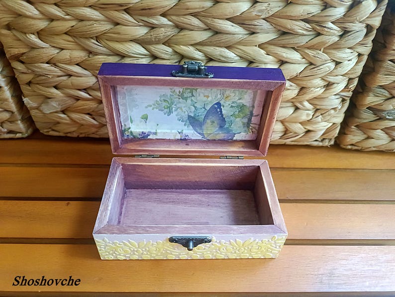 Puede incluir: Una caja de madera con una tapa morada y un dise&ntilde;o floral en el interior. La caja tiene un dise&ntilde;o de mariposa en el interior de la tapa y un dise&ntilde;o floral en el fondo. La caja est&aacute; abierta y vac&iacute;a.