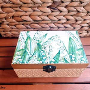 Puede incluir: Una pequeña caja de madera con una tapa con bisagras y un cierre de metal. La tapa está decorada con un patrón floral de flores blancas de lirio de los valles y hojas verdes sobre un fondo blanco. La caja tiene una base de color crema con un patrón tejido.