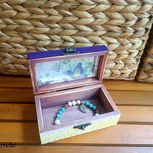 Puede incluir: Una caja de madera con una tapa morada y un dise&ntilde;o floral en el interior. La caja est&aacute; abierta y un brazalete de cuentas turquesa y marr&oacute;n con un dije de mariposa plateado est&aacute; en el interior.
