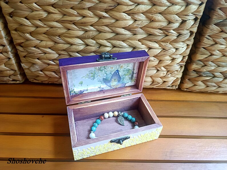 Puede incluir: Una caja de madera con una tapa morada y un dise&ntilde;o floral en el interior. La caja est&aacute; abierta y un brazalete de cuentas turquesa y marr&oacute;n con un dije de mariposa plateado est&aacute; en el interior.