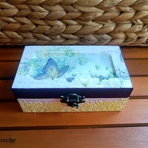 Puede incluir: Una peque&ntilde;a caja de madera con tapa abatible y cierre de metal. La caja est&aacute; decorada con un dise&ntilde;o floral y una mariposa. La caja est&aacute; sobre una superficie de madera.