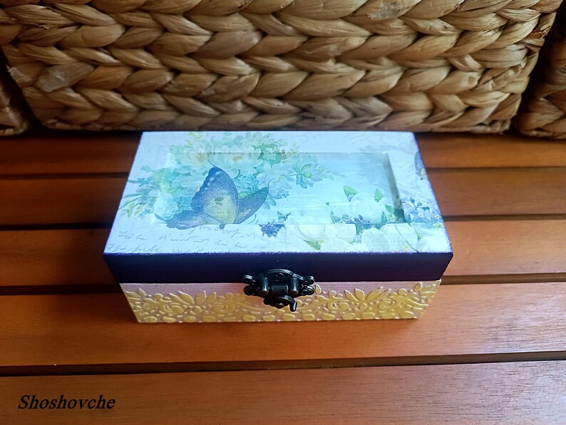 Puede incluir: Una peque&ntilde;a caja de madera con tapa abatible y cierre de metal. La caja est&aacute; decorada con un dise&ntilde;o floral y una mariposa. La caja est&aacute; sobre una superficie de madera.