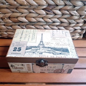 May include: A vintage-style wooden box with a hinged lid and a metal clasp. The box is decorated with a Parisian theme, featuring images of the Eiffel Tower, Notre Dame, and other landmarks. The box also has text in French, including "Visite la tres reelle Ville de Paris" and "Union des Commerçants de Sannois".