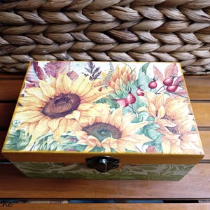 Puede incluir: Una caja de madera con tapa abatible y cierre de pestillo. La caja está decorada con un diseño floral colorido con girasoles, hojas y bayas rojas.