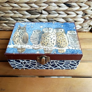 Peut inclure: Une boîte en bois avec un motif bleu et blanc représentant cinq hiboux perchés sur une branche. La boîte a un loquet et un couvercle brun foncé. Les hiboux sont représentés dans un style fantaisiste avec des étoiles et une lune en arrière-plan.