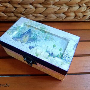 Puede incluir: Una peque&ntilde;a caja de madera con tapa con bisagras. La caja est&aacute; decorada con un dise&ntilde;o floral y una mariposa. La caja es de color azul y blanco.