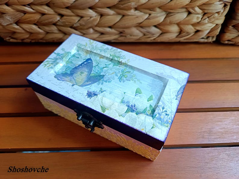 Puede incluir: Una peque&ntilde;a caja de madera con tapa con bisagras. La caja est&aacute; decorada con un dise&ntilde;o floral y una mariposa. La caja es de color azul y blanco.