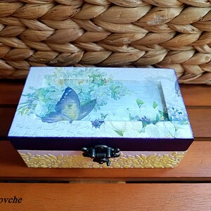 Puede incluir: Una peque&ntilde;a caja de madera con una tapa con bisagras y un cierre de metal. La caja est&aacute; decorada con un dise&ntilde;o floral y una mariposa azul. La caja est&aacute; sentada sobre una superficie de madera.