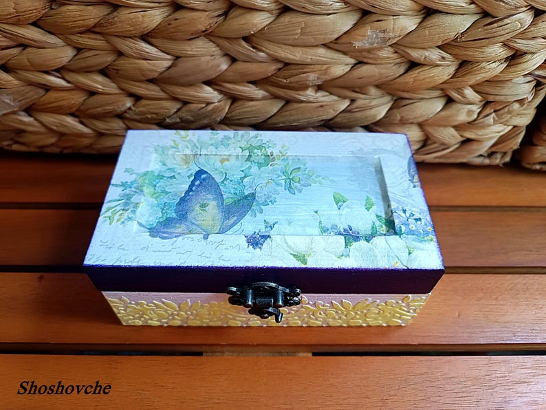 Puede incluir: Una peque&ntilde;a caja de madera con una tapa con bisagras y un cierre de metal. La caja est&aacute; decorada con un dise&ntilde;o floral y una mariposa azul. La caja est&aacute; sentada sobre una superficie de madera.