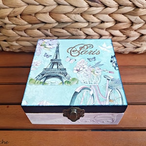 Puede incluir: Una caja de madera con fondo azul claro y borde negro. La caja presenta una imagen pintada de la Torre Eiffel, una bicicleta con una cesta de flores y la palabra "París" en letras doradas brillantes.
