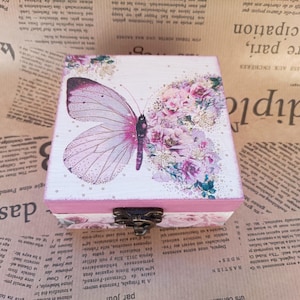 May include: A decorative wooden box with a butterfly and floral design. The box features a hinged lid and a metal clasp. The butterfly is pink and purple, and the flowers are pink and purple. The box is on a newspaper background.